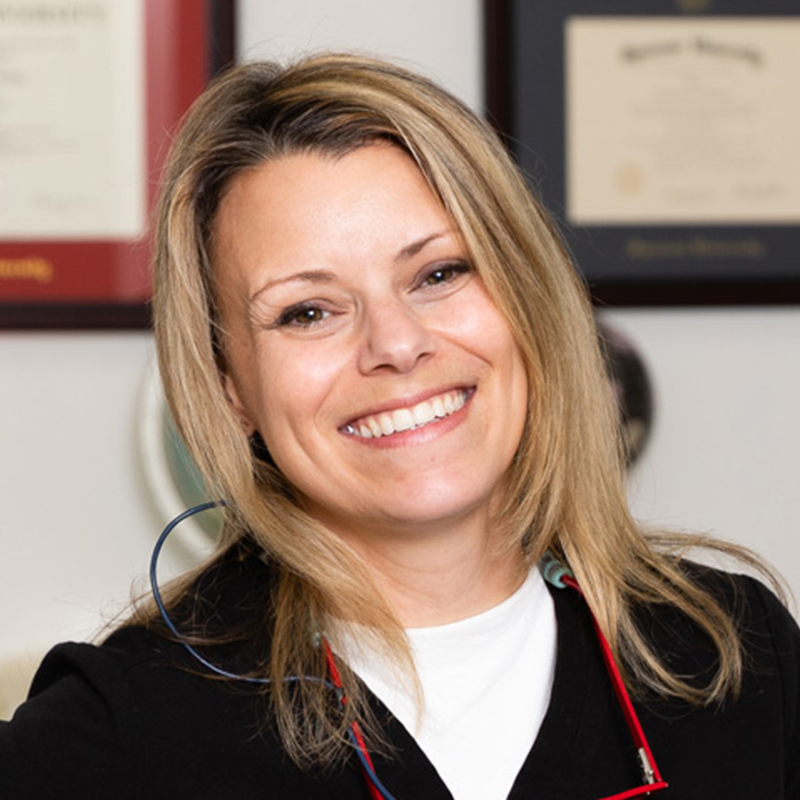A smiling woman with long, light brown hair and glasses, standing in an office setting adorned with framed certificates, exuding a friendly and professional demeanor.