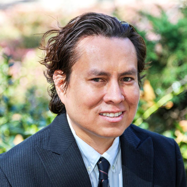A man with medium-length, wavy hair smiles confidently while wearing a black suit and tie, with lush greenery in the background.