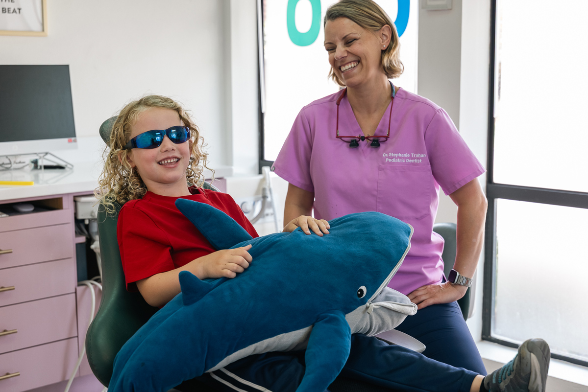 A smiling child wearing sunglasses holds a plush shark while sitting in a dental chair, accompanied by a cheerful dentist in a pink shirt.