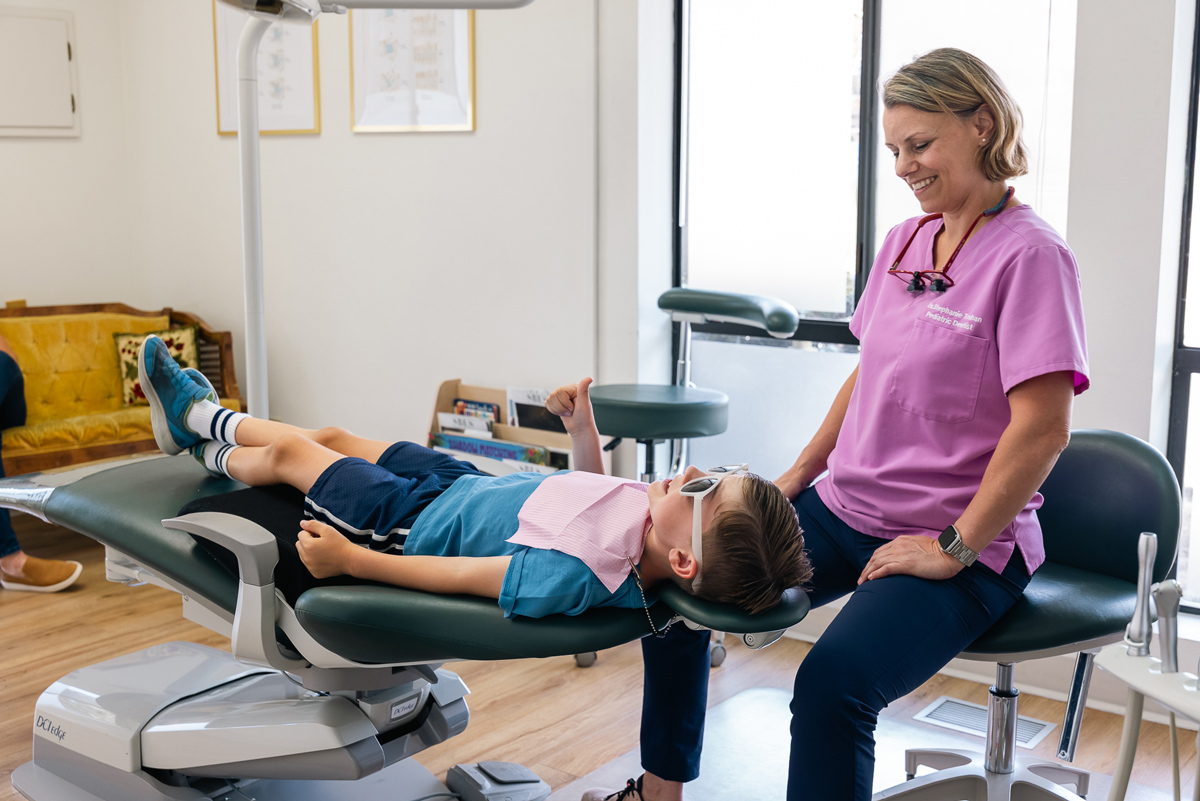 A healthcare professional is assisting a young patient lying on a dental chair, both appearing relaxed in a well-lit office setting.