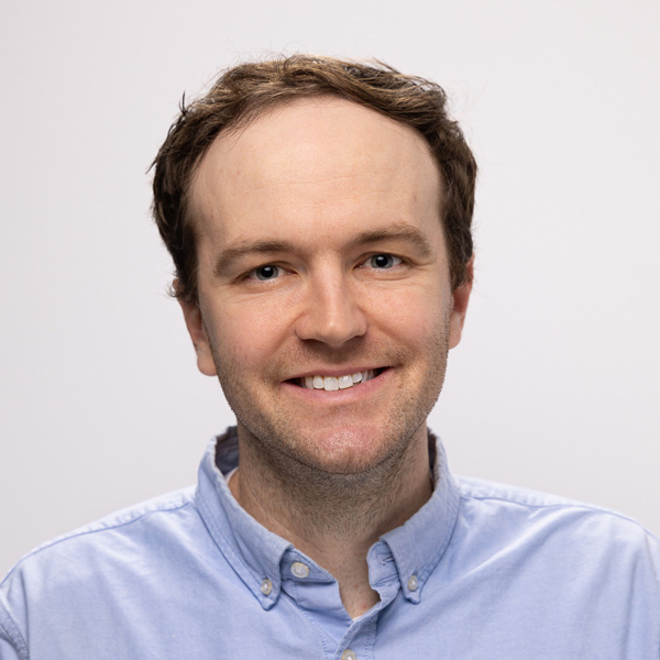 A smiling individual with light brown hair is wearing a blue button-up shirt. The background is plain and neutral, creating a friendly and professional vibe.